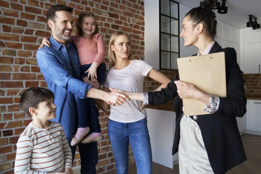 Family shaking hands with real estate agent in new apartments 4 women gathered around a desk smiling and reviewing blueprints